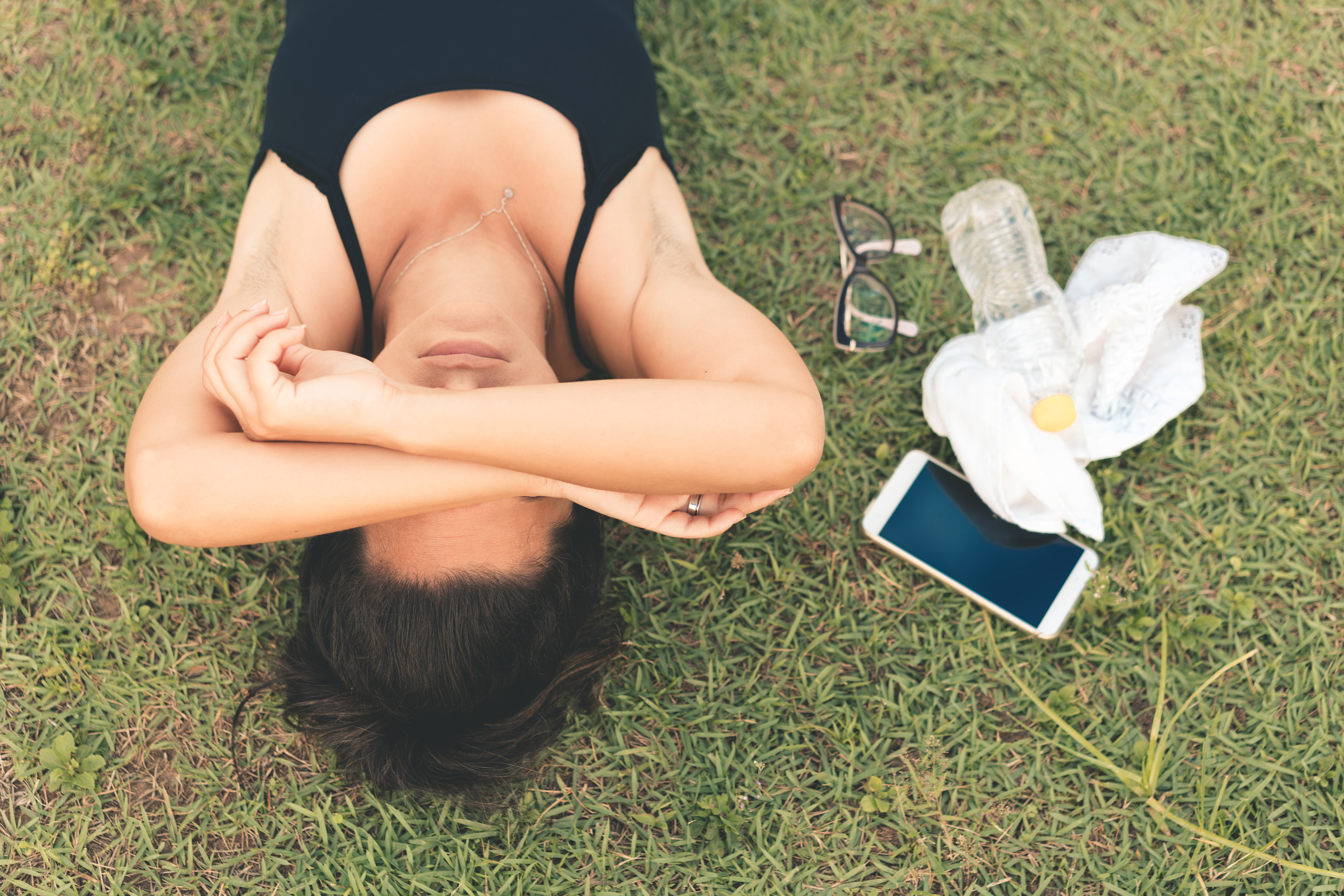 A woman lying on the grass with her arms crossed over her face lying next to a water bottle and towel