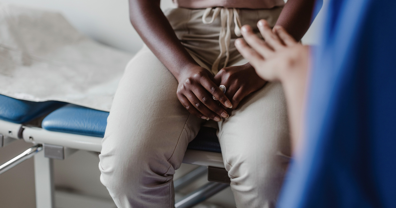 A patient sitting on an exam table with her hands in her lap listening to a doctor