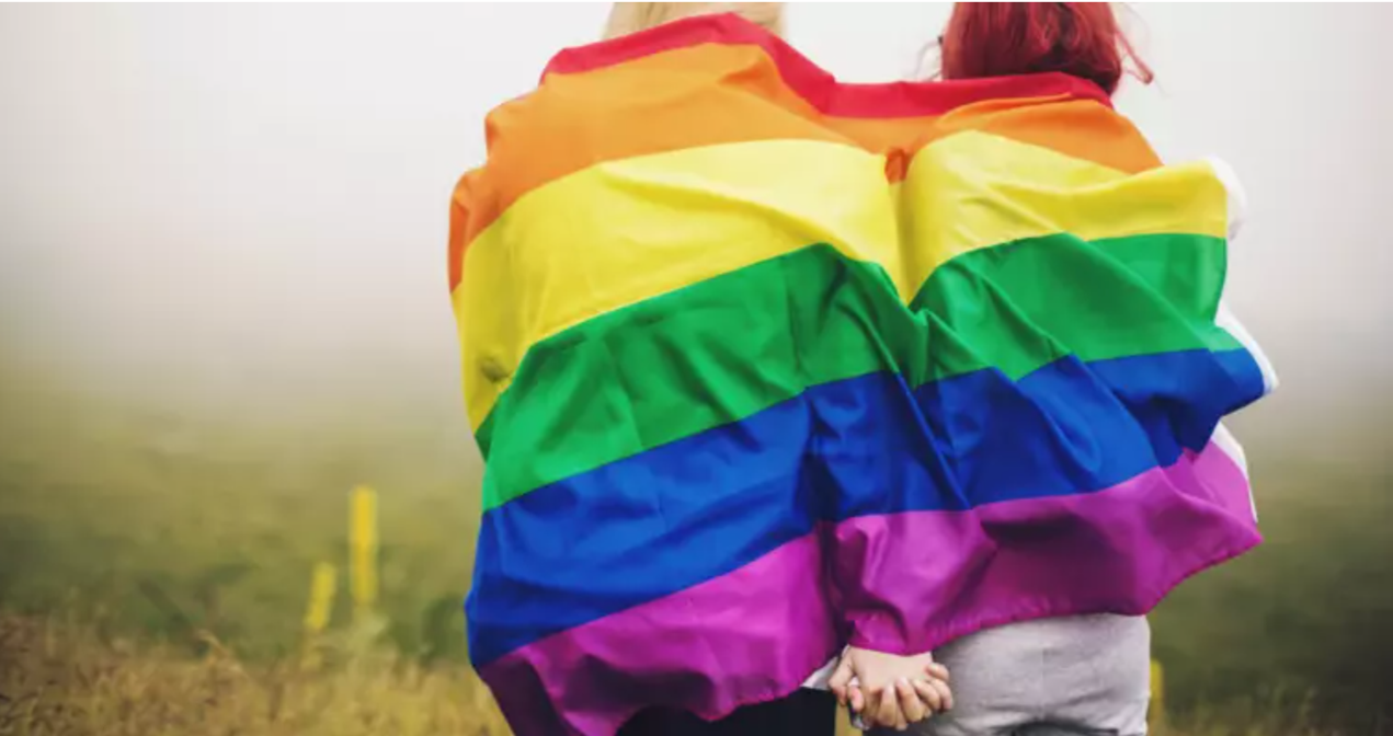 The back of a young couple holding hands with a pride flag draped around their shoulders