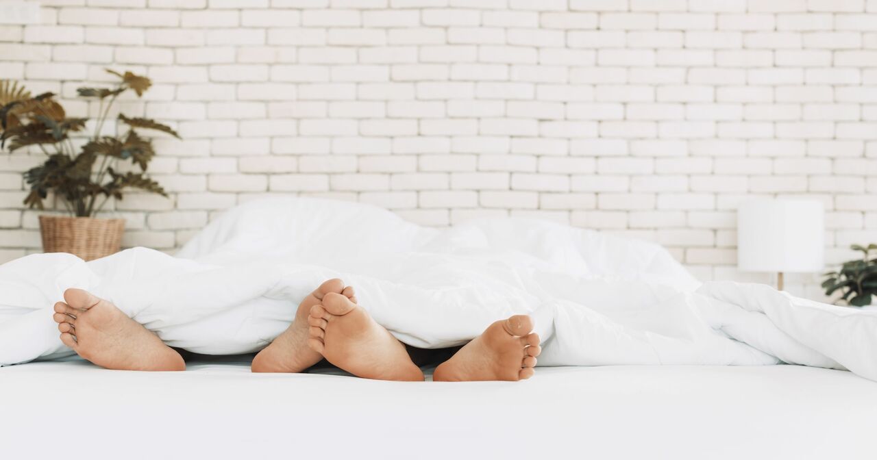 The bare feet of two couples lying side by side in a bed with white bedding