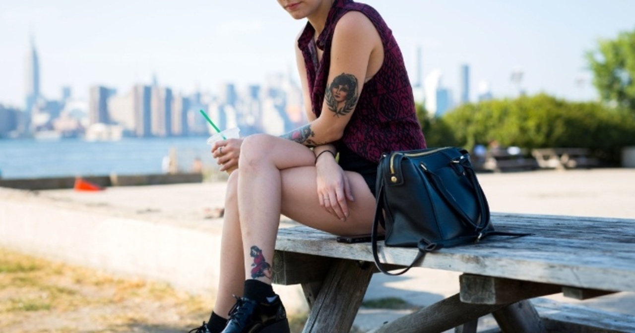 A young woman with tattoos sitting on top of a picnic table with the city skyline behind her